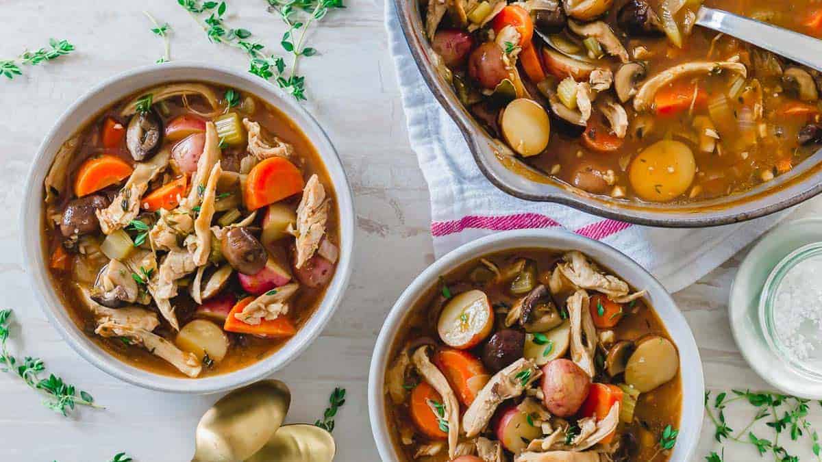 Three bowls of chicken stew with carrots, potatoes, mushrooms, and celery, garnished with herbs, are arranged on a white table next to a pot and gold spoons.