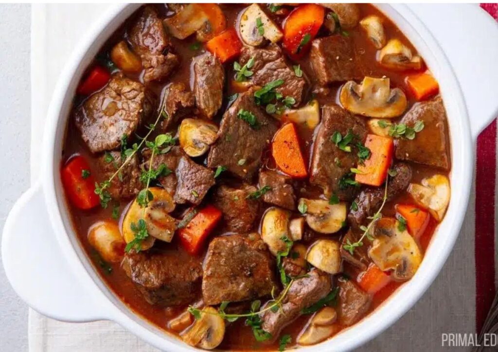 A bowl of beef stew with chunks of beef, sliced mushrooms, carrots, and sprigs of fresh herbs in a rich brown broth.
