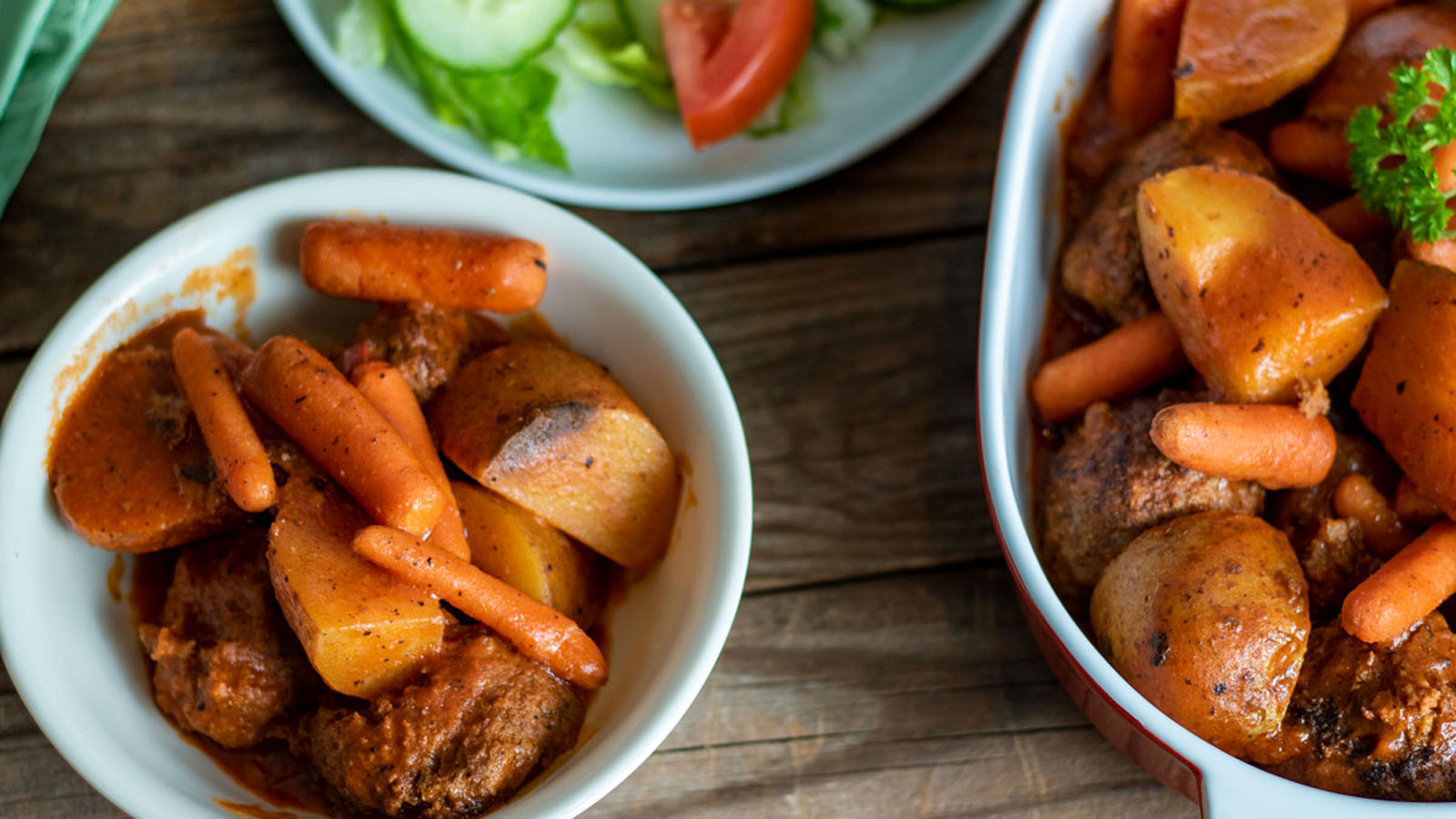 Bowls of beef stew with potatoes and carrots in a reddish-brown sauce, served alongside a plate of sliced cucumber, lettuce, and tomato on a wooden surface.