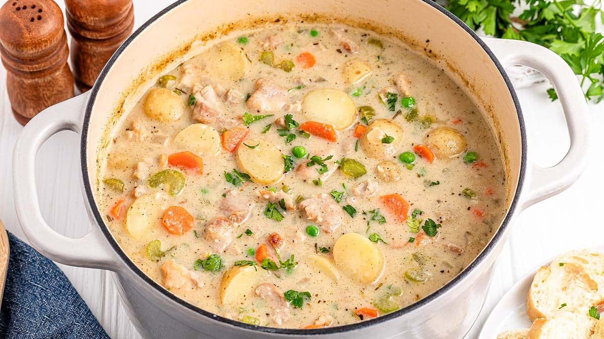 A pot of creamy chicken stew with sliced potatoes, carrots, peas, and herbs, sits on a white surface next to salt and pepper shakers.
