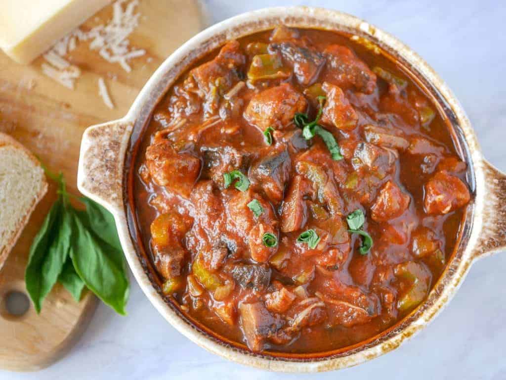 A bowl of tomato-based stew with chunks of meat and vegetables, garnished with fresh herbs. A cutting board with bread, cheese, and basil is nearby.