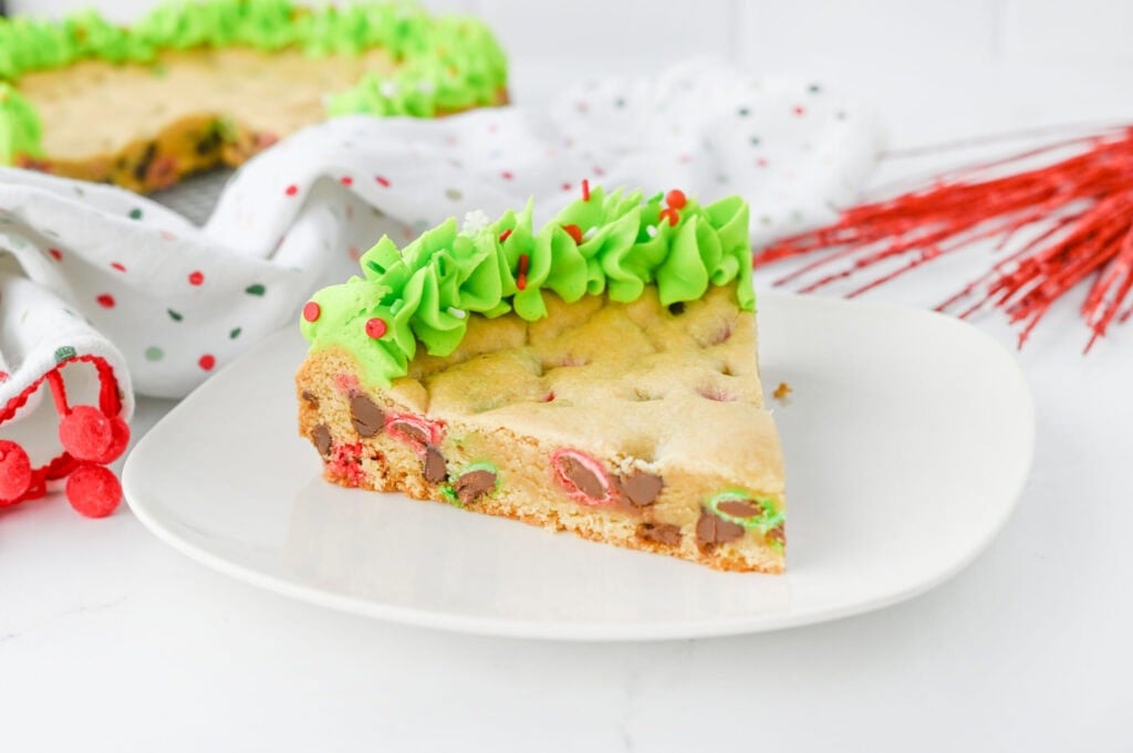 A slice of cookie cake with green frosting and red sprinkles sits on a white plate, with a decorated napkin and more cookie cake in the background.