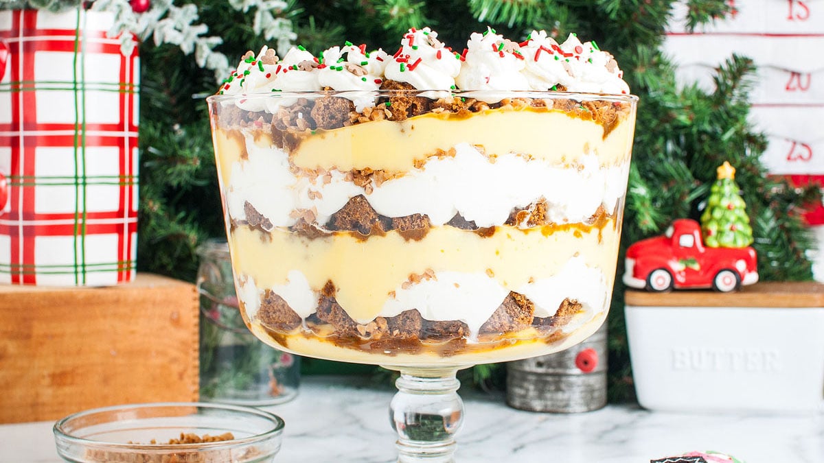 A layered trifle dessert with brownie pieces, whipped cream, and pudding in a glass bowl, topped with whipped cream and holiday sprinkles, set in a festive Christmas setting.