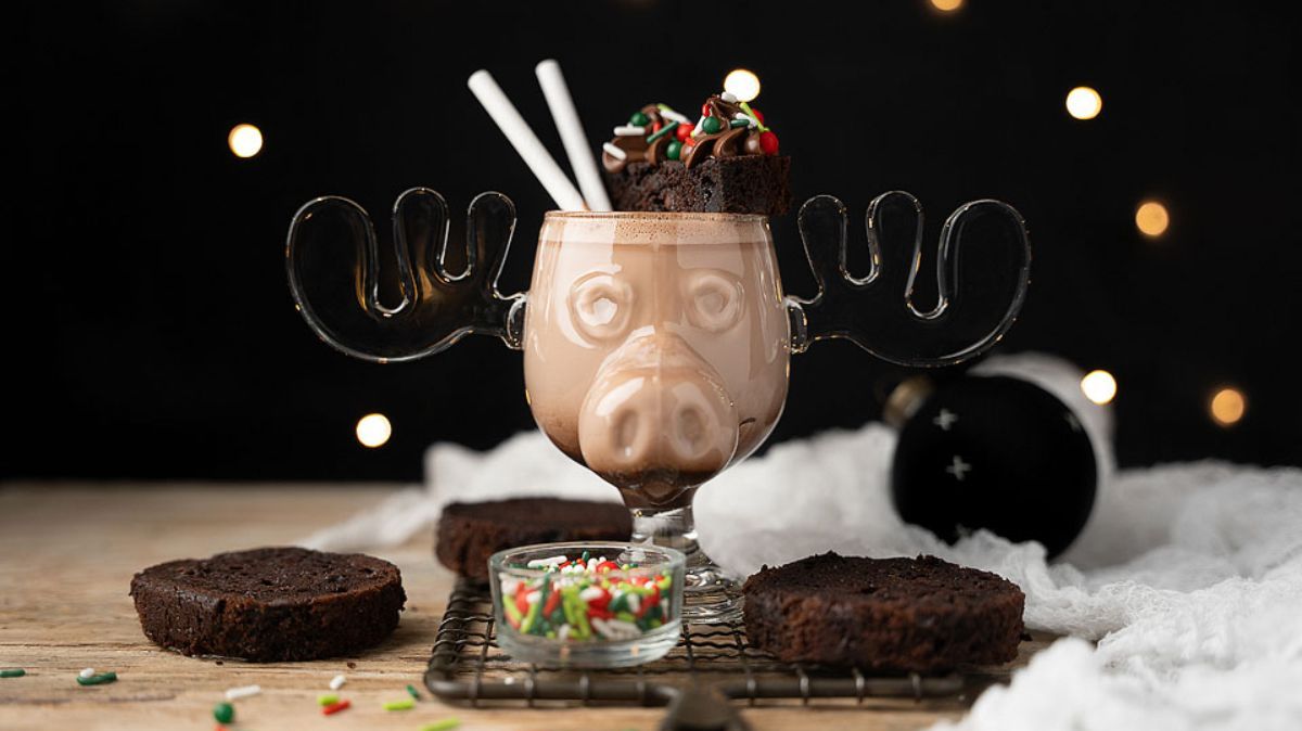 A glass shaped like a moose head filled with chocolate milk, topped with brownies and sprinkles, surrounded by slices of chocolate cake and a small bowl of sprinkles.