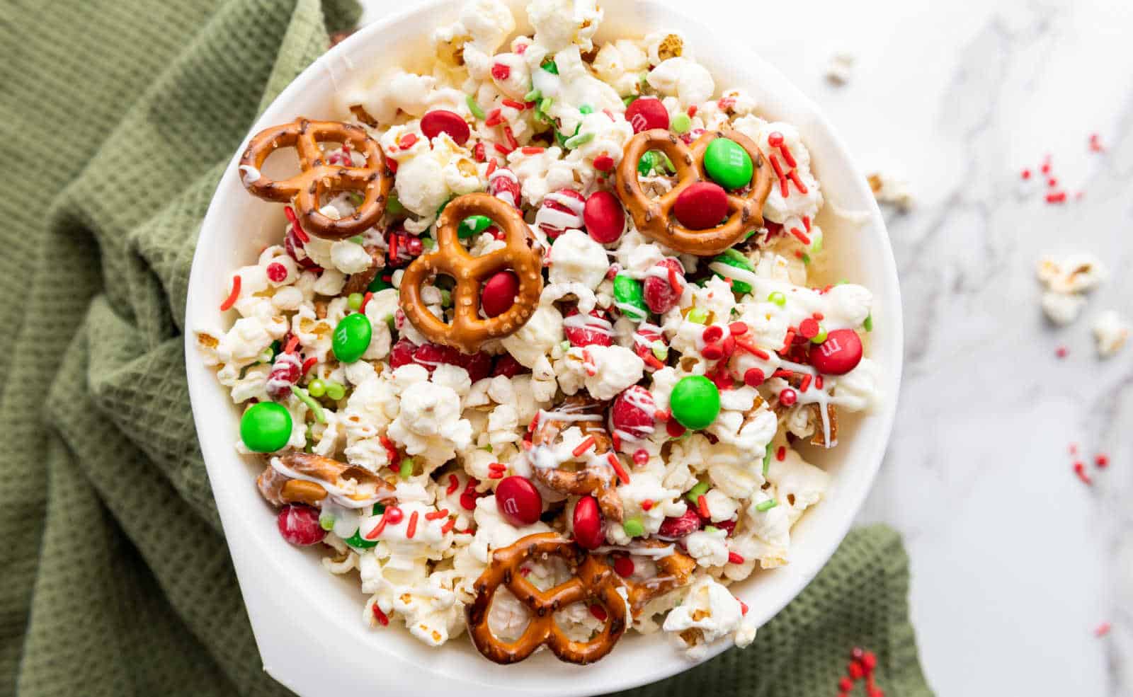 A bowl of popcorn mixed with red and green candies, pretzels, and red sprinkles, placed on a green textured cloth.