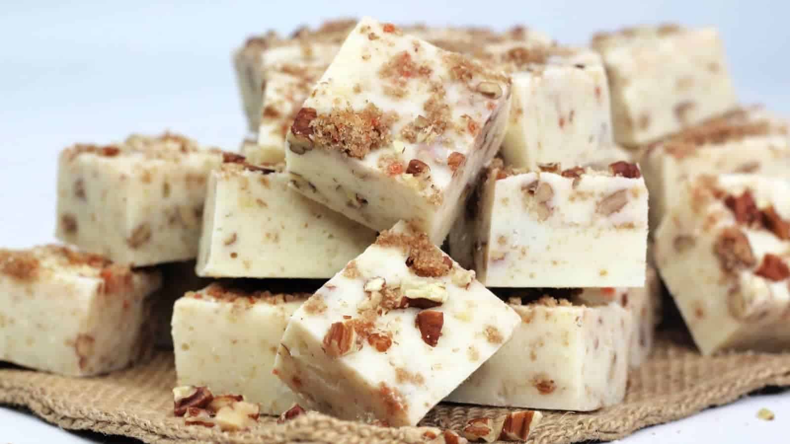 Squares of white nut fudge are stacked on a burlap cloth, with visible pieces of chopped nuts mixed throughout and sprinkled on top.