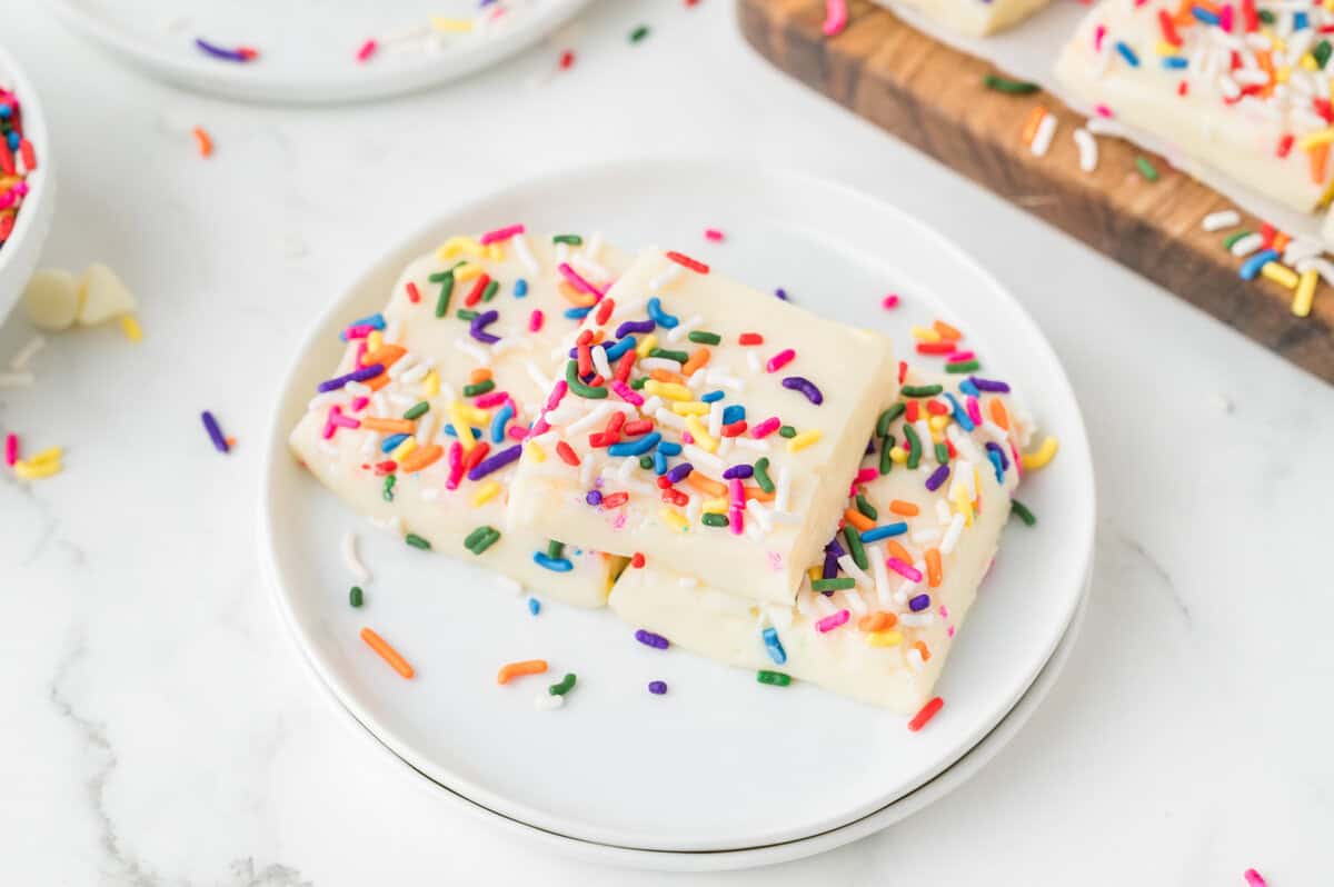 Two rectangular pieces of white fudge topped with colorful rainbow sprinkles are stacked on a white plate.