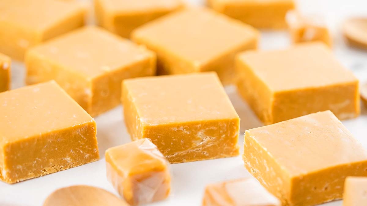Several pieces of square, golden brown caramel fudge are arranged on a white surface, with one piece partially wrapped in clear plastic.