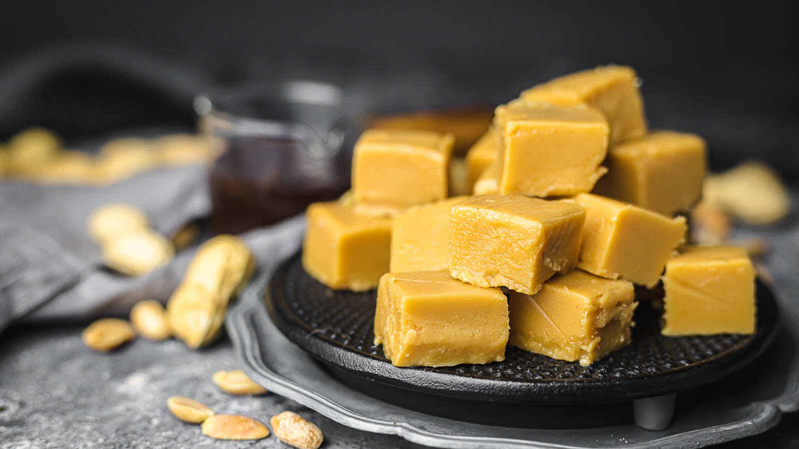 A stack of square peanut butter fudge pieces arranged on a black plate, with peanuts scattered nearby and a glass jar in the background.