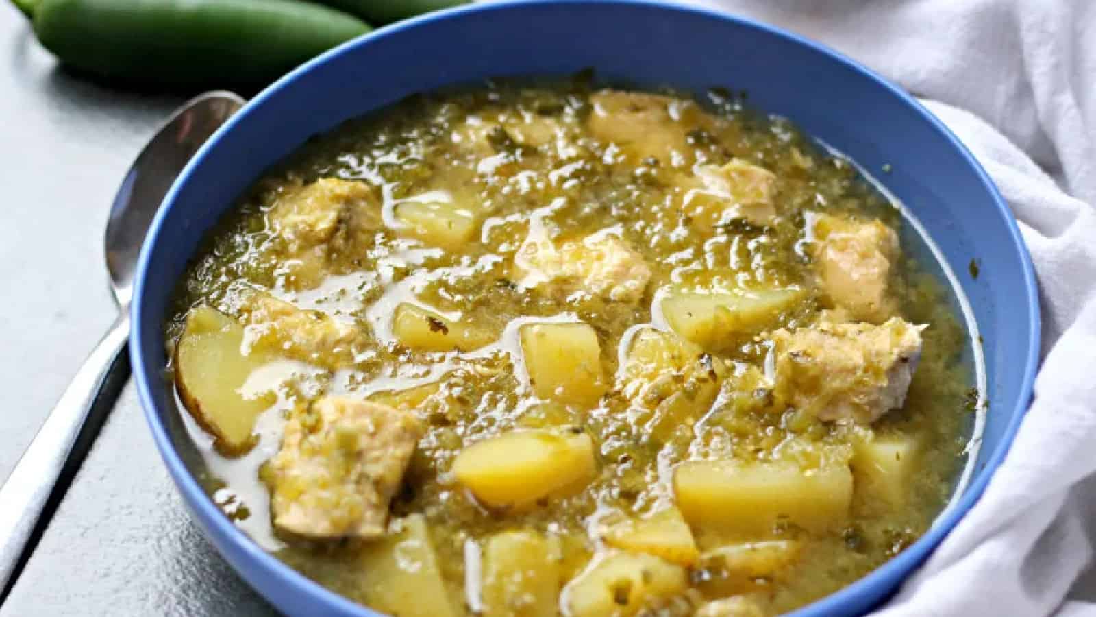 A blue bowl filled with green chile stew containing chunks of potato and meat, placed beside a spoon and a green chili pepper.