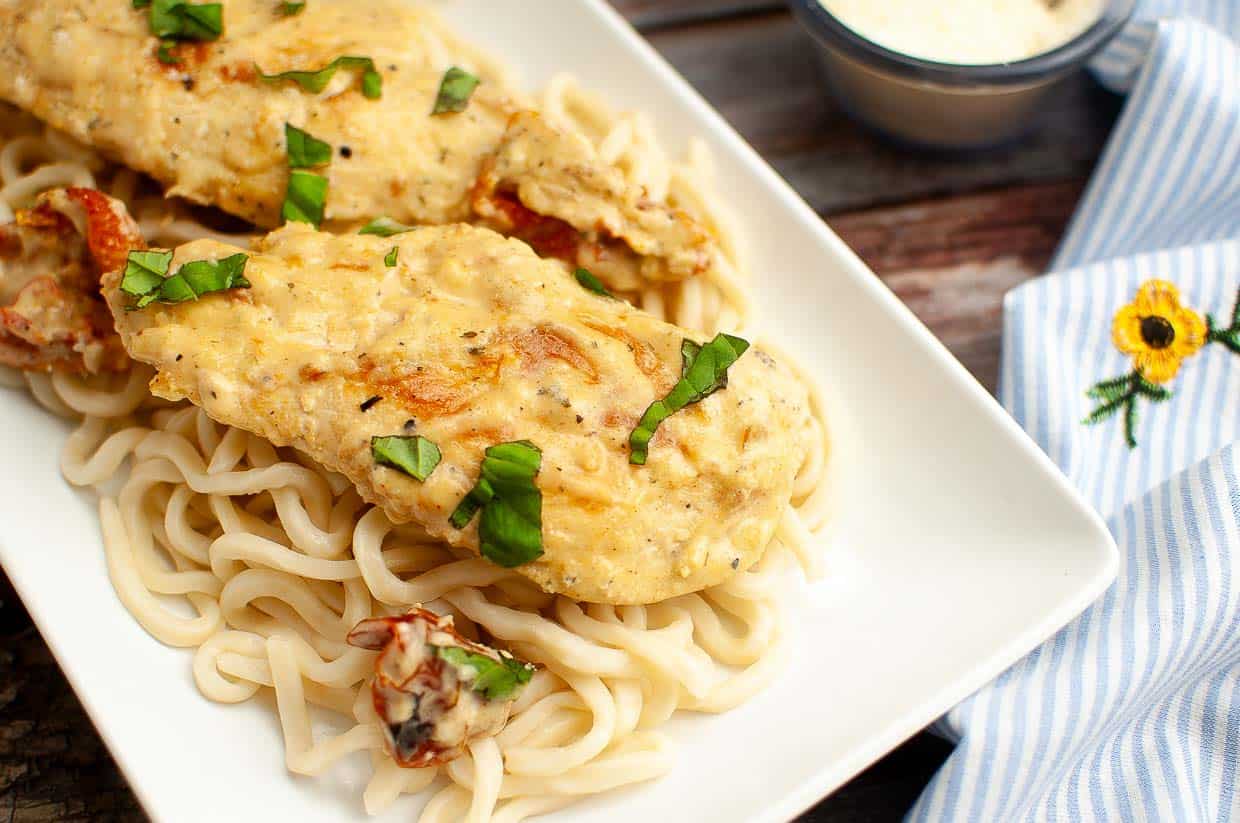 Chicken breasts in creamy sauce served over a bed of pasta, garnished with chopped basil on a white rectangular plate.