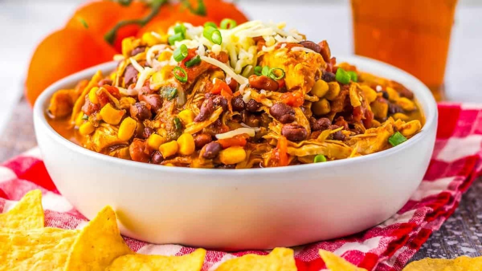 A bowl of chicken chili with beans, corn, shredded cheese, and green onions, placed on a red checkered cloth next to tortilla chips and fresh tomatoes.