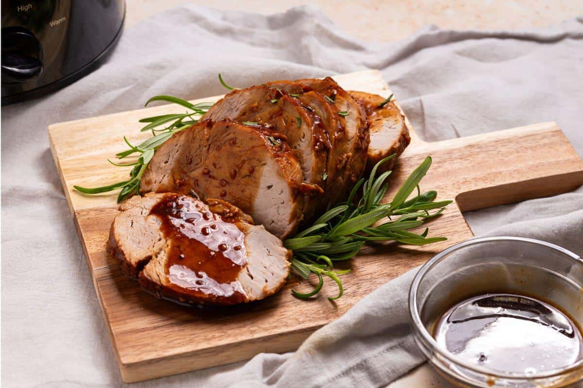 Sliced roast pork loin with sauce and sprigs of rosemary on a wooden cutting board, next to a bowl of sauce.
