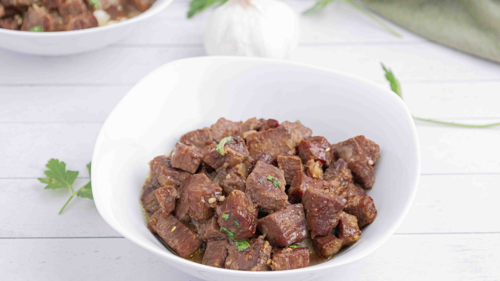 A white bowl filled with cooked, cubed beef garnished with parsley, placed on a light-colored surface with garlic and herbs in the background.