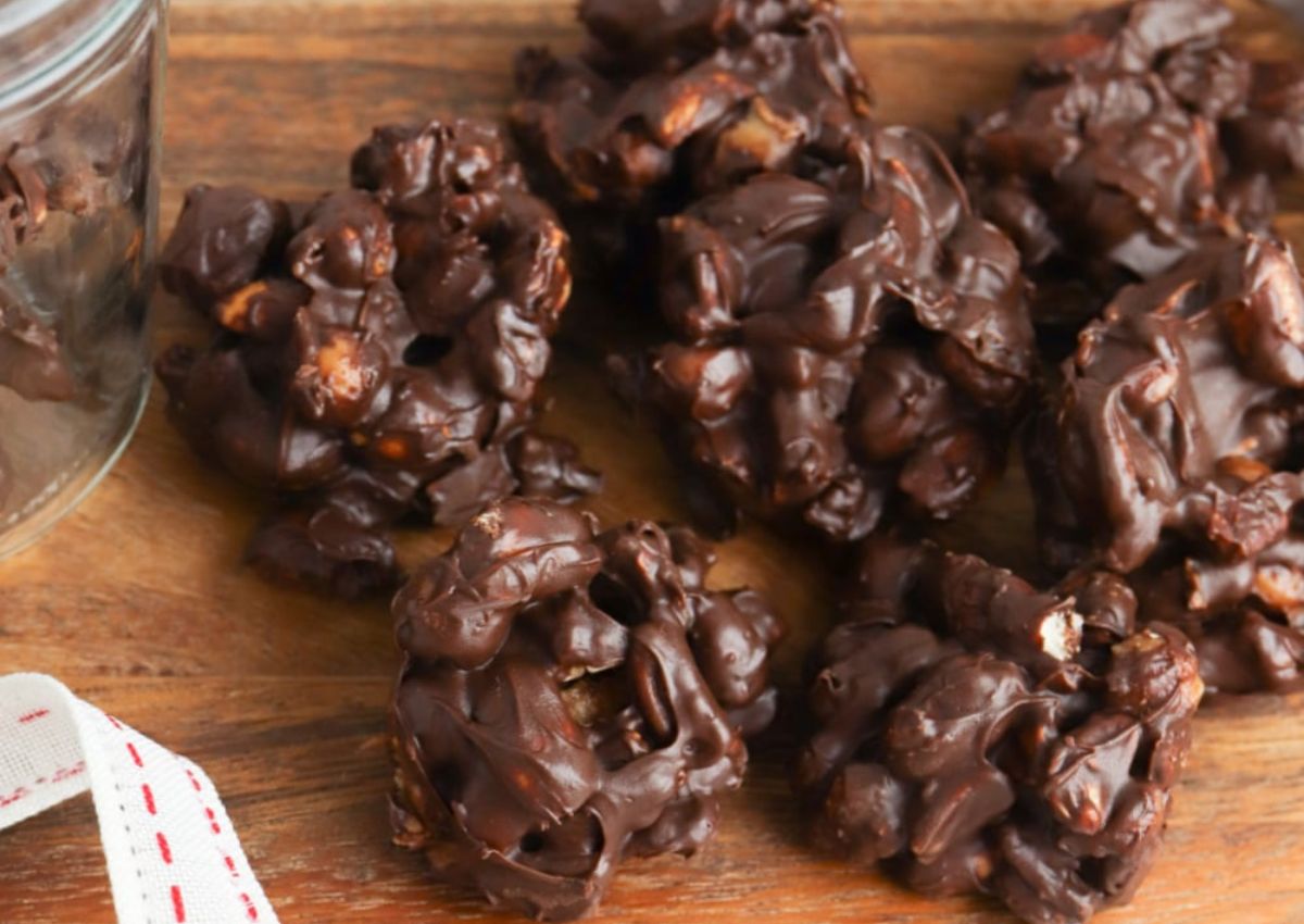 Clusters of chocolate-covered nuts are arranged on a wooden surface next to a glass jar.