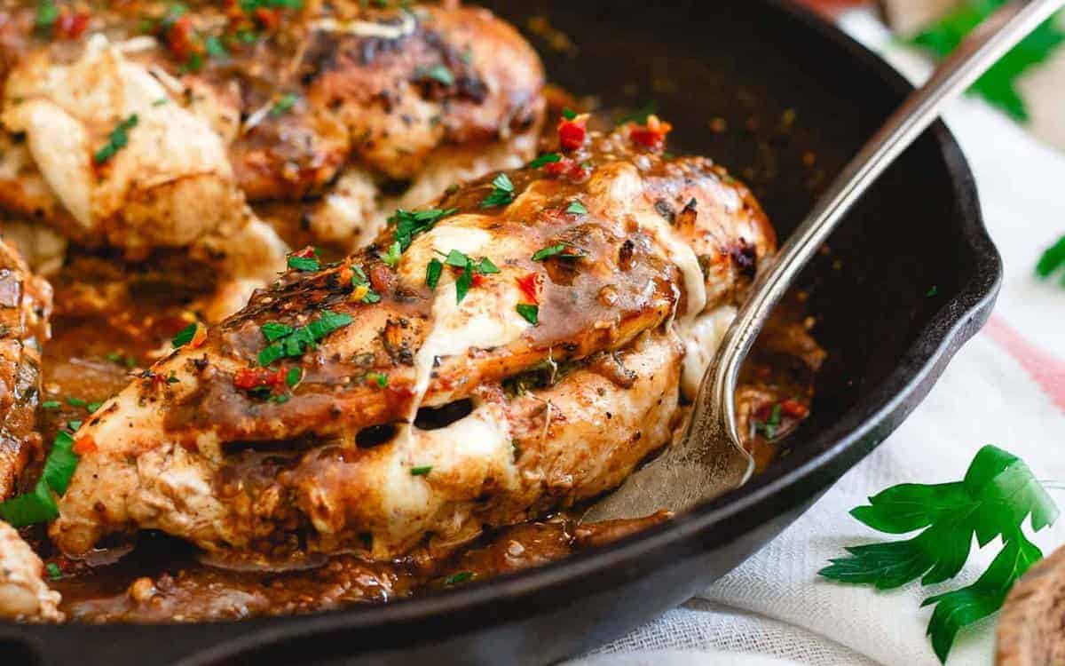 Cooked chicken breast stuffed with melted cheese and herbs, garnished with chopped parsley, in a black skillet with a serving spoon.