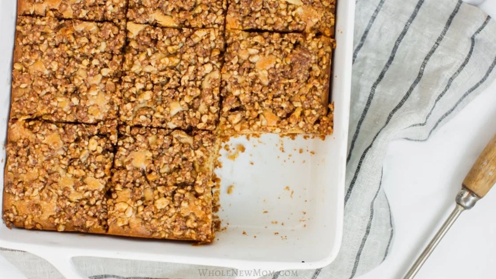A white baking dish of crumb-topped coffee cake cut into squares, with one piece missing. A striped towel and a serving utensil are beside the dish.