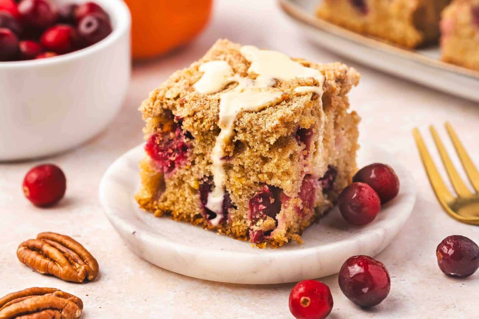 A slice of cranberry crumb cake topped with glaze sits on a small plate, surrounded by fresh cranberries and pecans, with a fork and bowl of cranberries nearby.