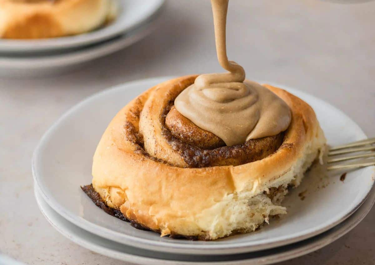 A close-up of a cinnamon roll on a white plate being topped with a swirl of brown icing.
