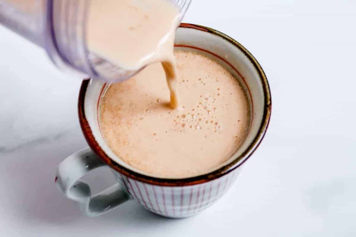 A creamy light-brown beverage is being poured from a blender into a white mug with red stripes, placed on a white surface.
