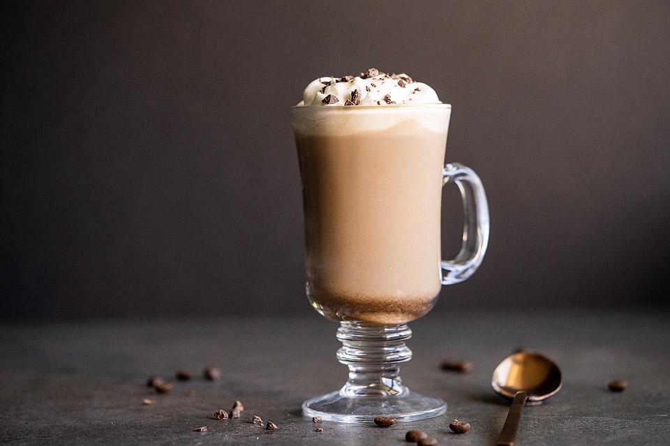 A glass mug filled with a creamy coffee beverage topped with whipped cream and chocolate shavings, with coffee beans and a spoon beside it on a dark surface.
