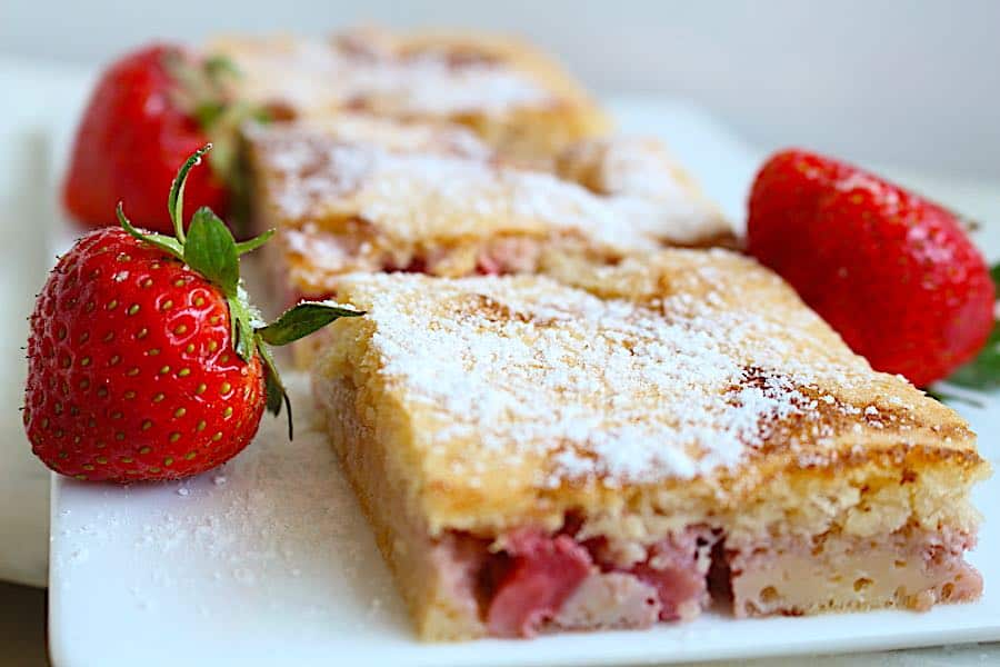 Three rectangular slices of strawberry cake dusted with powdered sugar are arranged on a white plate, with fresh strawberries placed nearby.