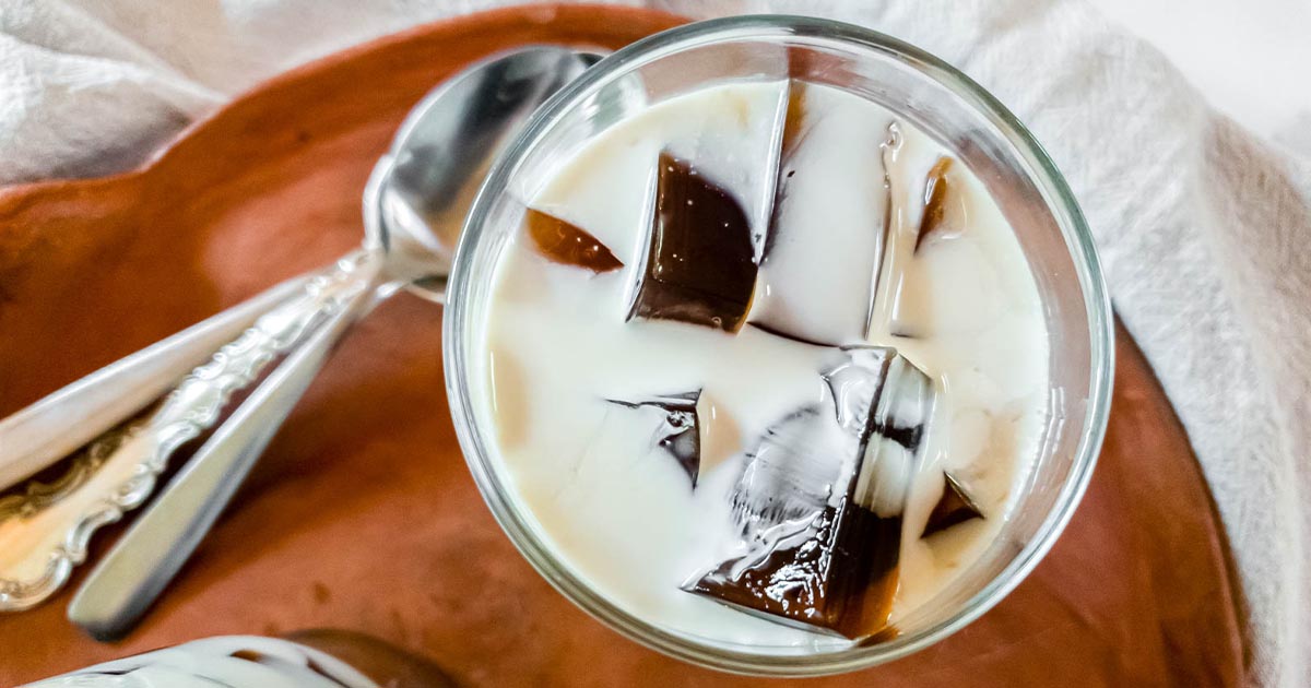 Glass filled with a creamy white beverage and dark ice cubes, placed on a brown surface next to two metal spoons.