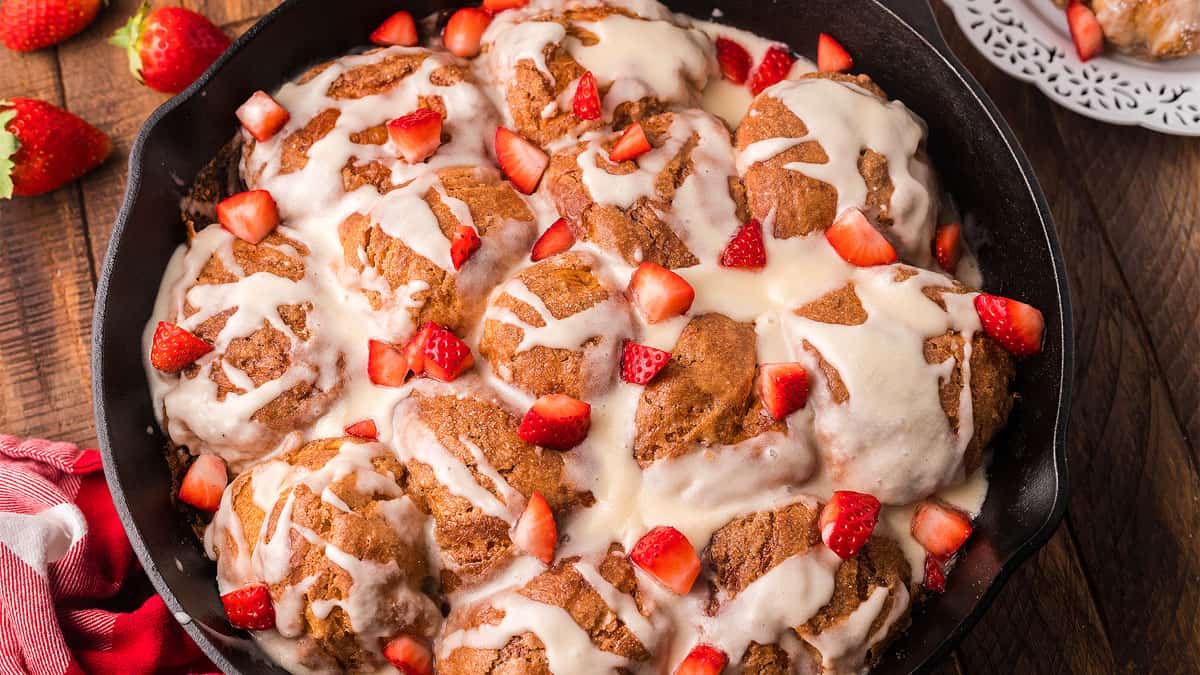 Skillet of cinnamon rolls topped with white icing and fresh strawberry pieces, placed on a wooden table with whole strawberries nearby.