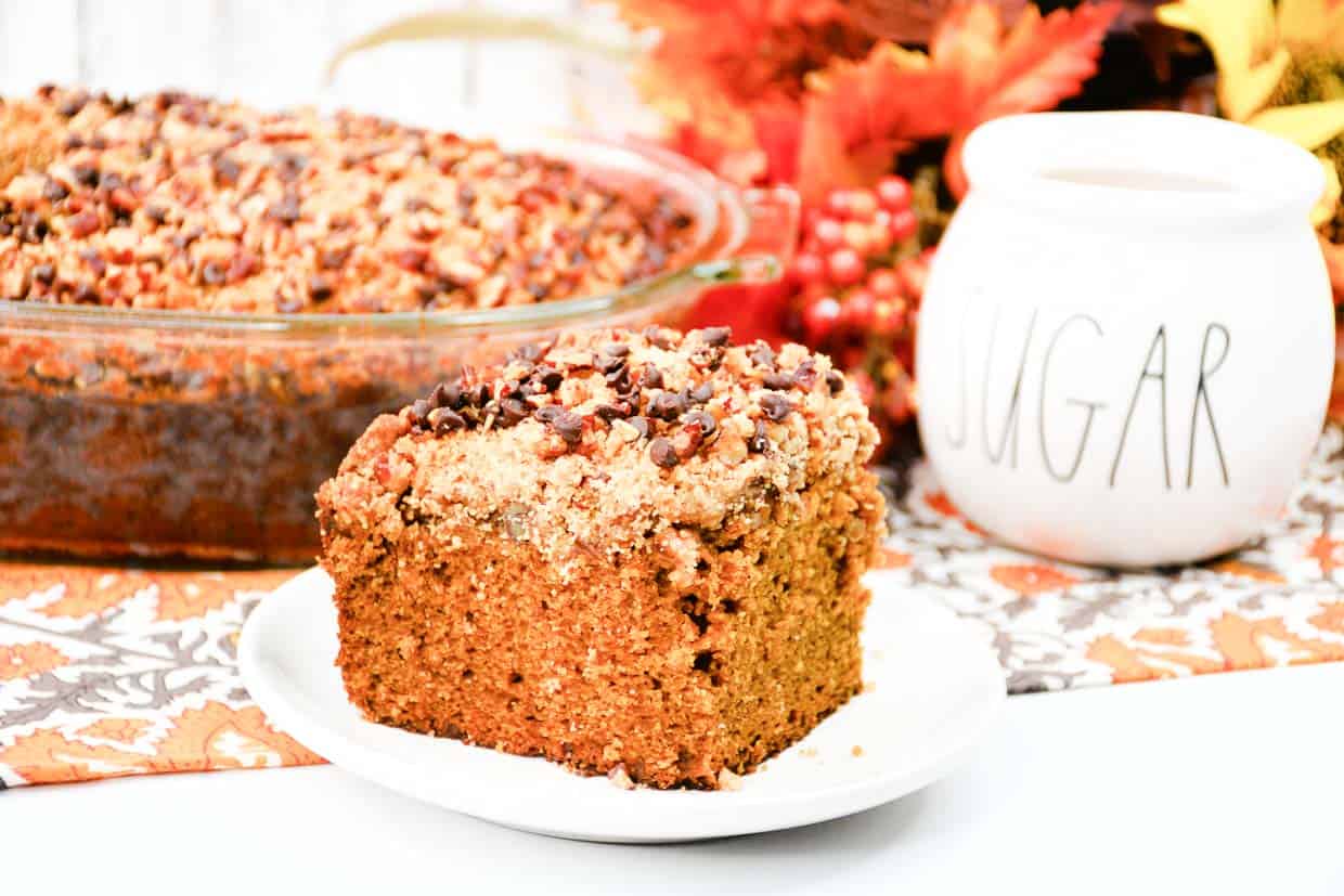 A slice of crumb-topped coffee cake sits on a white plate in front of a baking dish, with a SUGAR jar and fall decorations in the background.
