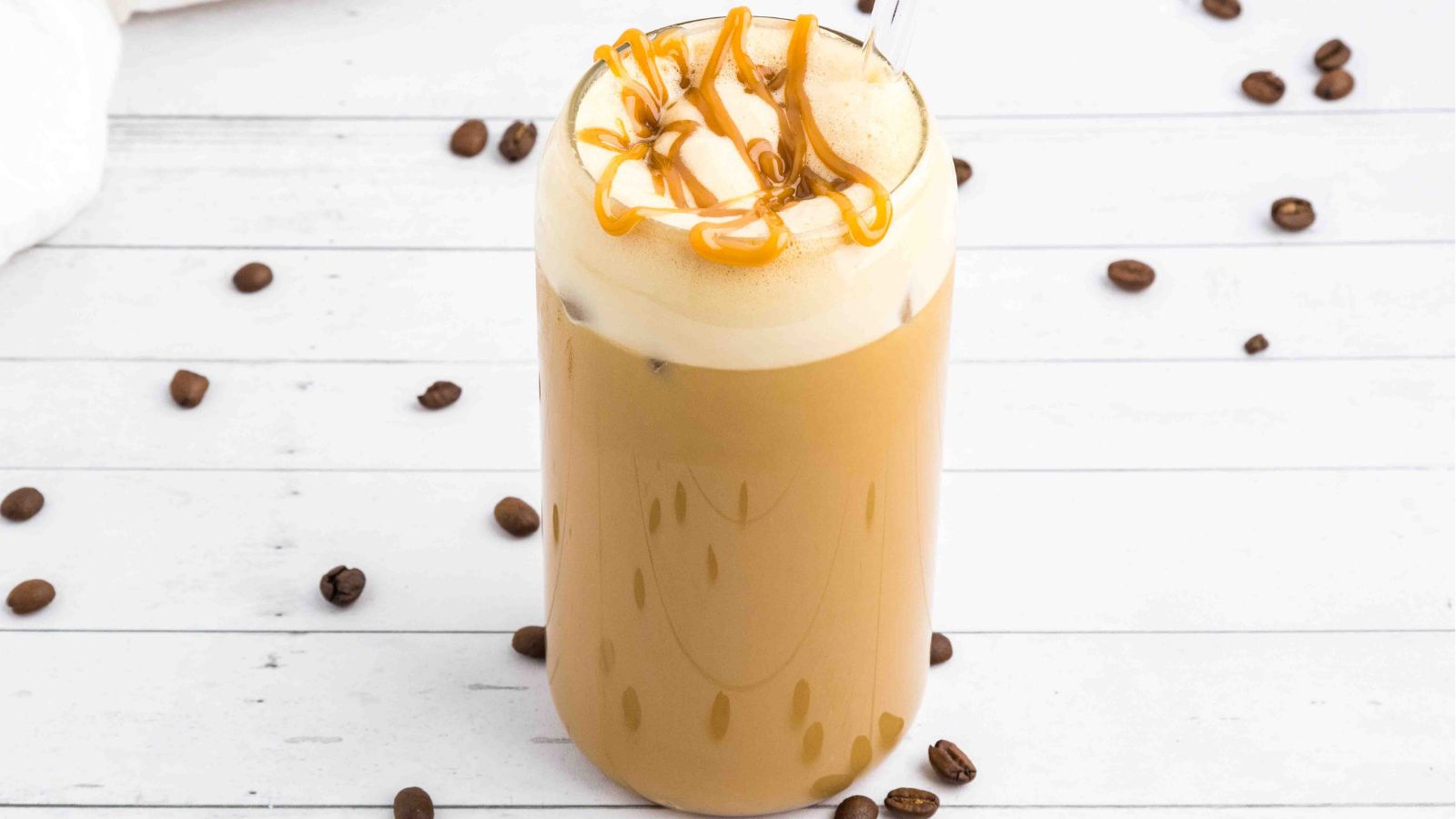 A glass of iced coffee topped with whipped cream and caramel drizzle, with a straw, sits on a white wooden surface scattered with coffee beans.
