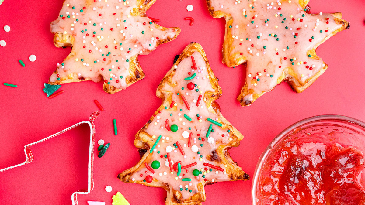 Three Christmas tree-shaped cookies with pink icing and colorful sprinkles are on a red surface, next to a bowl of red jam and a metal cookie cutter.