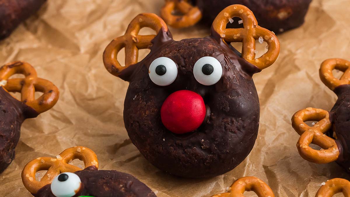 A chocolate-covered donut decorated with candy eyes, a red candy nose, and pretzels for antlers, resembling a reindeer, sits on brown parchment paper.