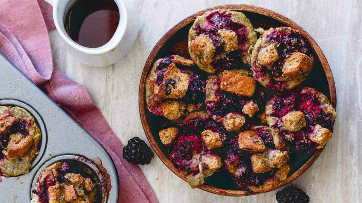 A wooden bowl filled with blackberry bread pudding muffins sits on a table next to a muffin tin, a pink cloth, a cup of coffee, and a few fresh blackberries.