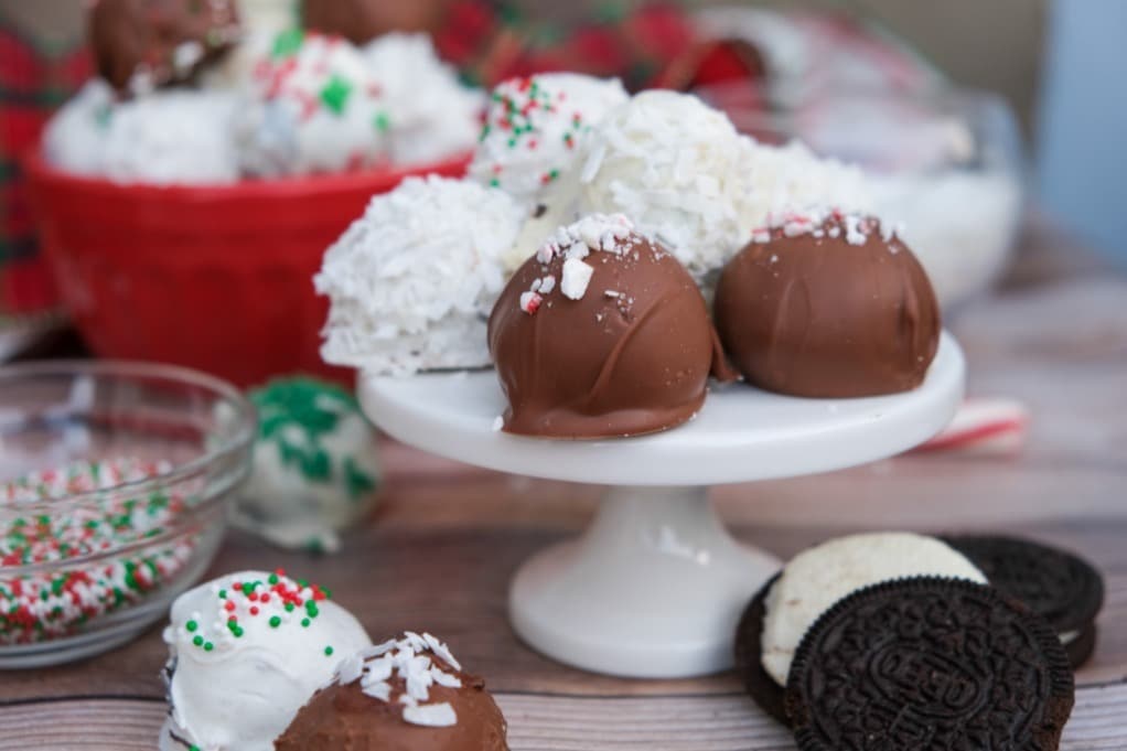 An assortment of chocolate and white-coated truffles with sprinkles and coconut shreds displayed on a small cake stand, with Oreo cookies and bowls of sprinkles nearby.