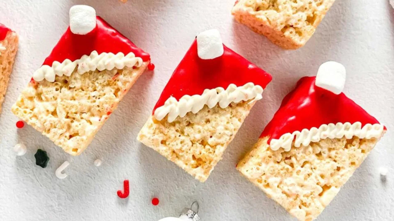 Rice Krispies treats decorated with red icing, white piped frosting, and mini marshmallows to resemble Santa hats, arranged on a white surface.