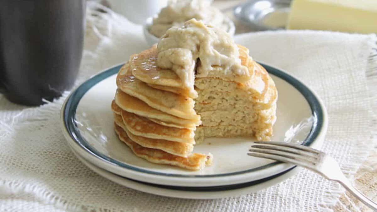 A stack of pancakes with a dollop of cream on top sits on a plate; a bite has been cut out, and a fork rests beside the pancakes.