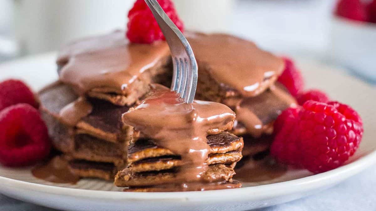 A stack of pancakes covered in chocolate sauce, garnished with fresh raspberries, with a fork cutting into the stack.