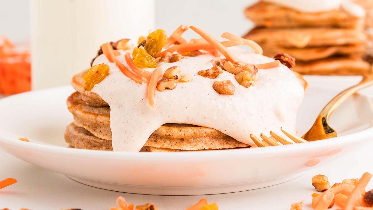 A stack of pancakes topped with cream, shredded carrots, golden raisins, and chopped walnuts on a white plate with a gold fork.
