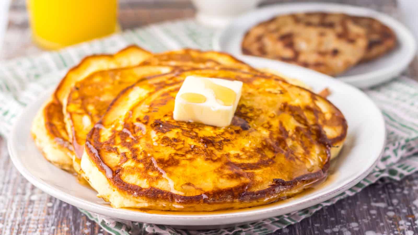 A plate of pancakes topped with a pat of butter and syrup, with a glass of orange juice and another dish in the background.