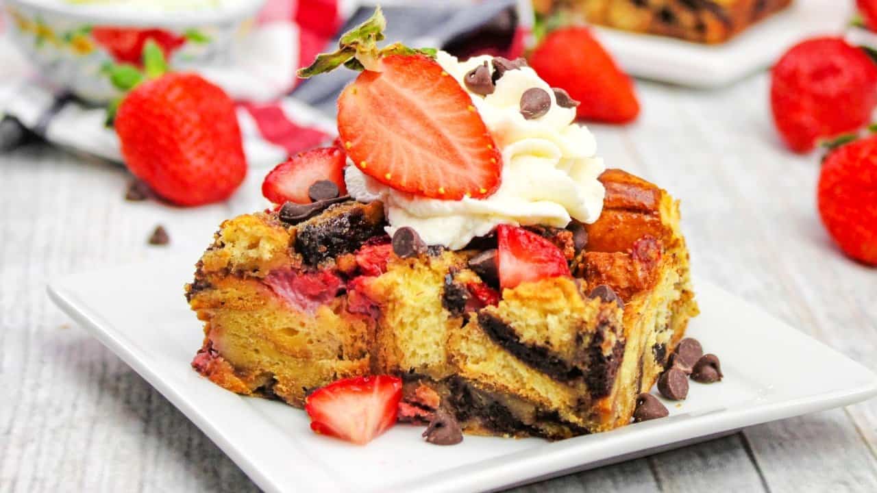 A slice of bread pudding with strawberries, chocolate chips, and whipped cream on top, served on a white plate with whole strawberries in the background.
