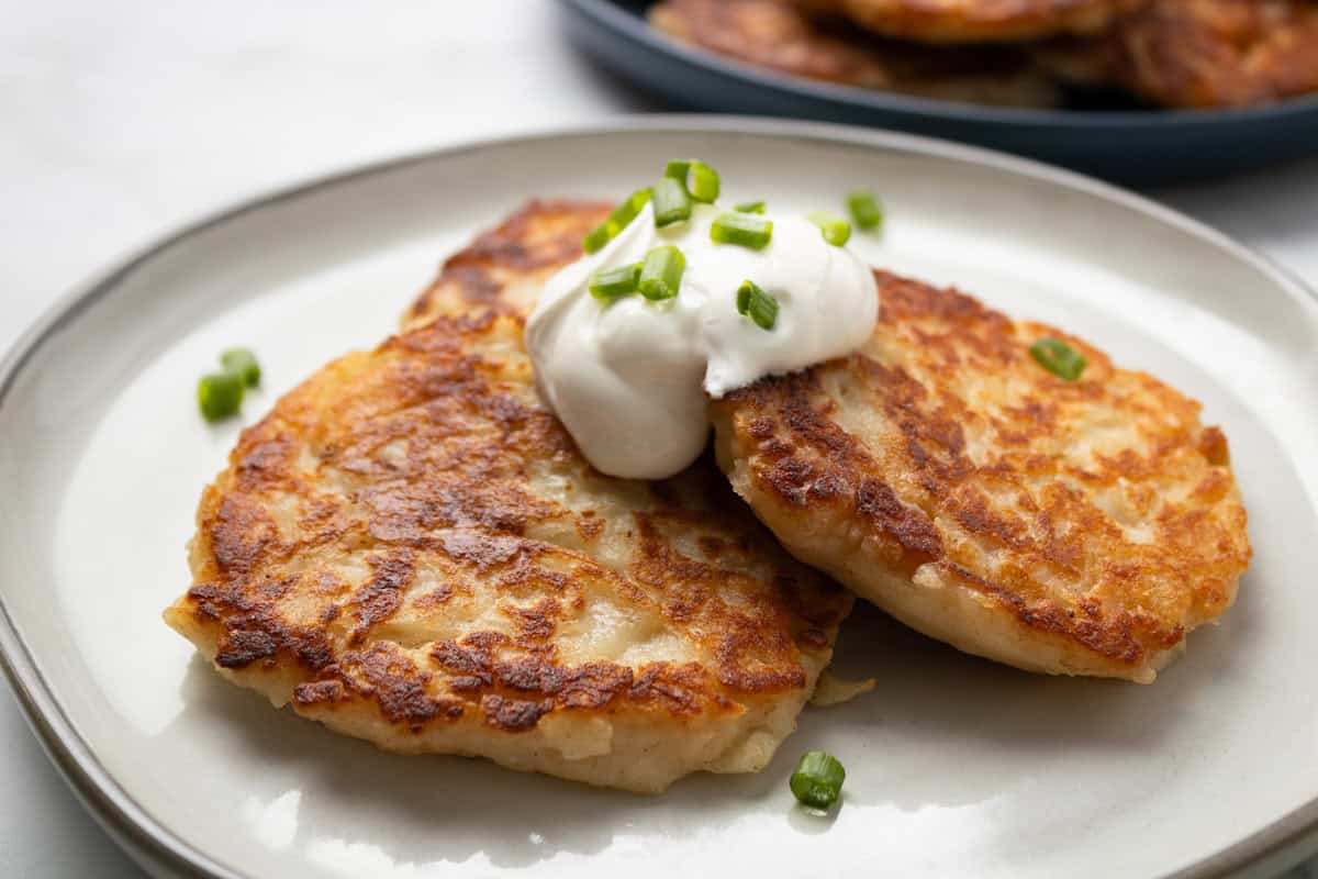 Two golden-brown potato pancakes topped with a dollop of sour cream and chopped green onions on a white plate.