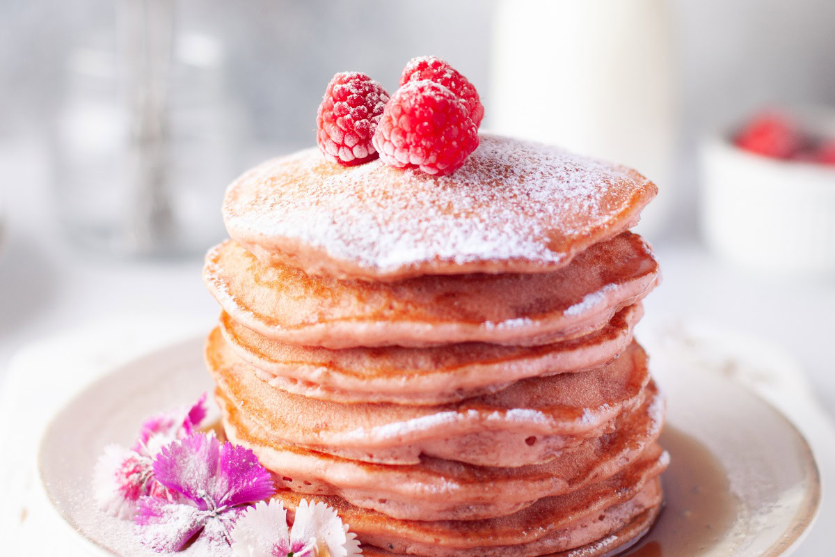 A stack of pink pancakes topped with powdered sugar and raspberries, garnished with edible flowers on a white plate.