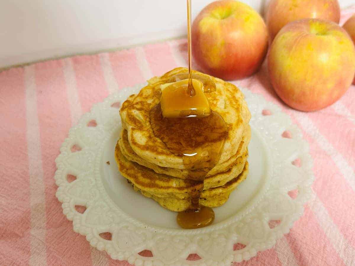 A stack of pancakes topped with a pat of butter and syrup being poured, on a white plate with three apples in the background.