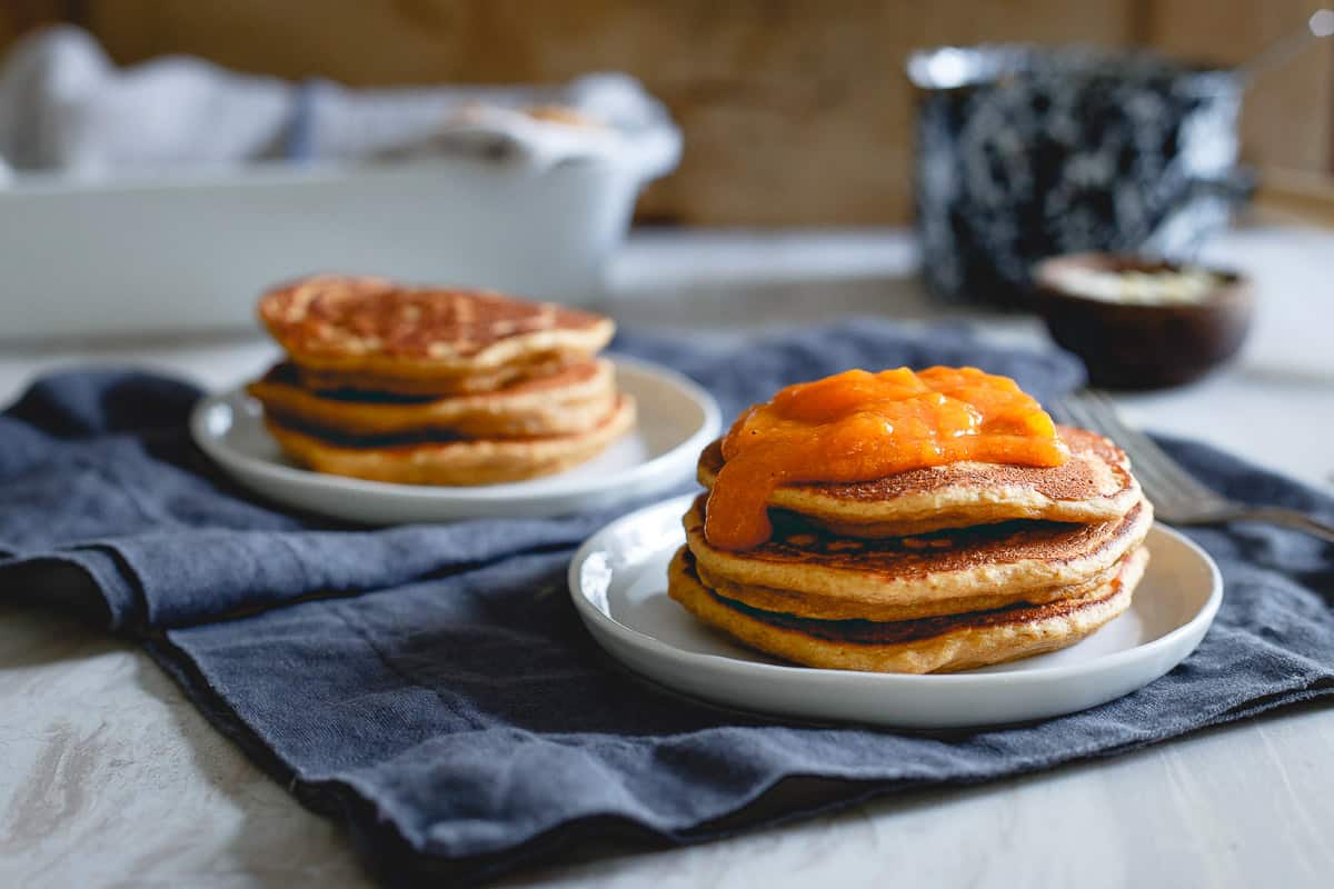Two small stacks of pancakes sit on white plates; one stack is topped with a bright orange fruit sauce. Both are placed on a dark cloth over a light table.