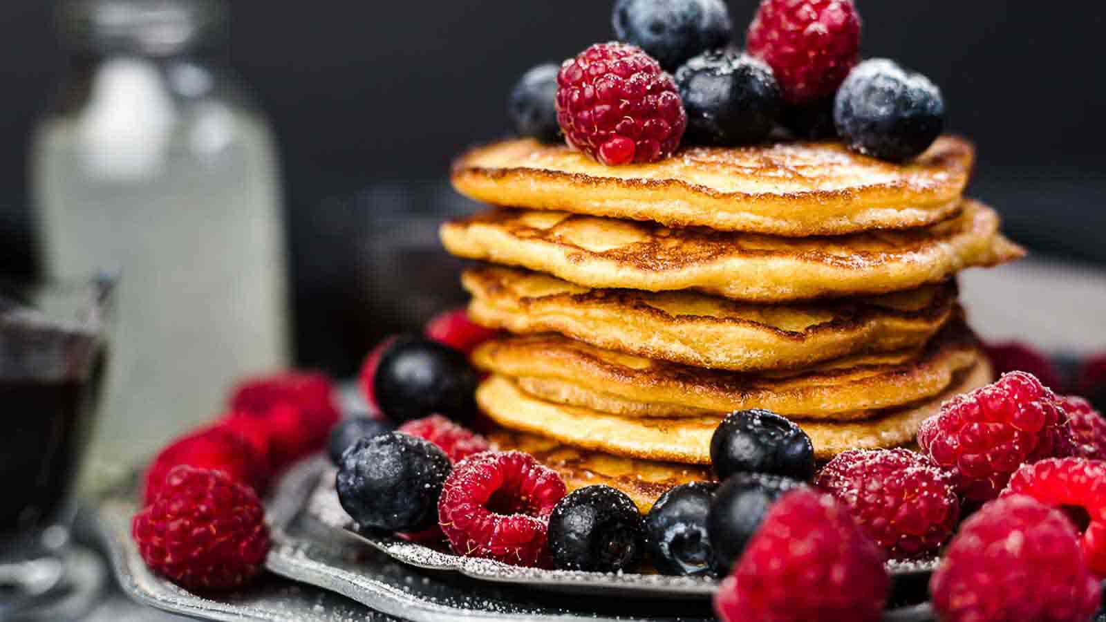 A stack of pancakes topped with fresh raspberries and blueberries, with more berries scattered around on a plate.