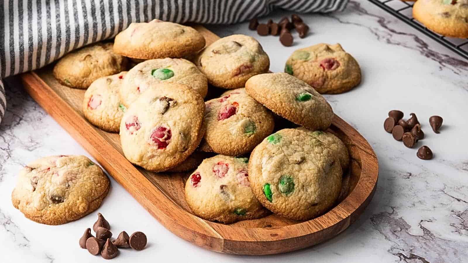 A wooden tray filled with cookies containing red and green candies and chocolate chips, with extra chocolate chips scattered nearby.