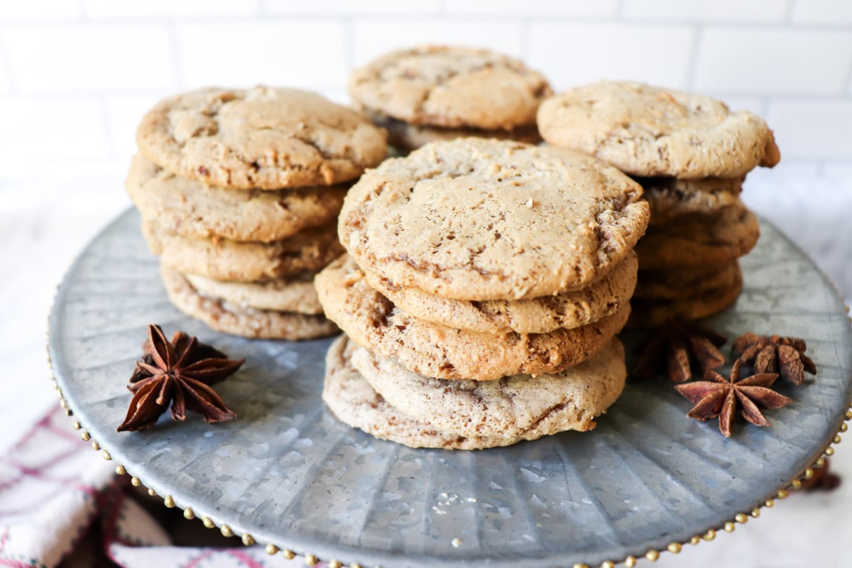 Eight stacks of round cookies are arranged on a metal cake stand, with star anise pods placed nearby. White tile background.