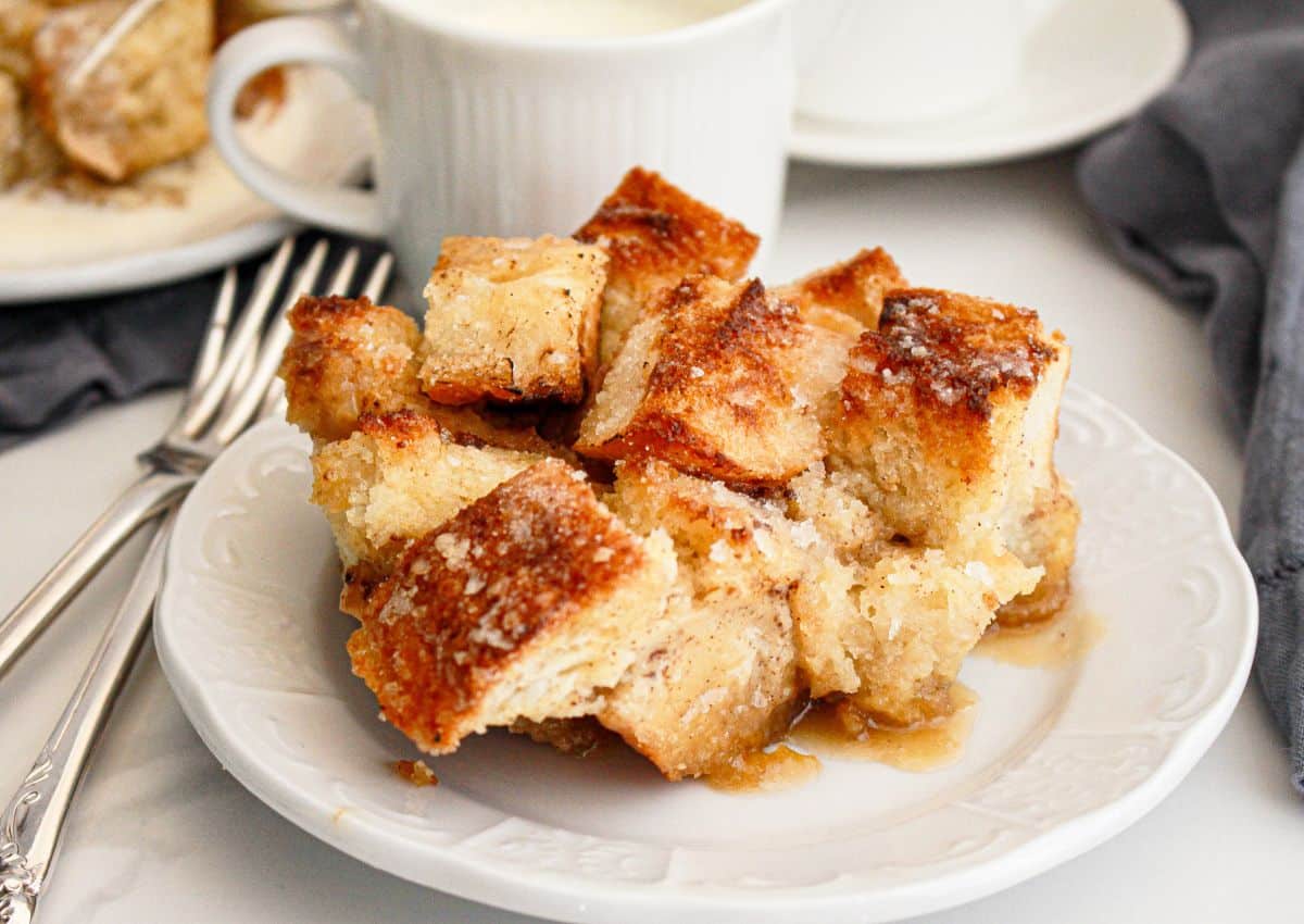 A serving of bread pudding with a golden, crispy top is plated on a white dish, with forks, a cup, and a napkin nearby.