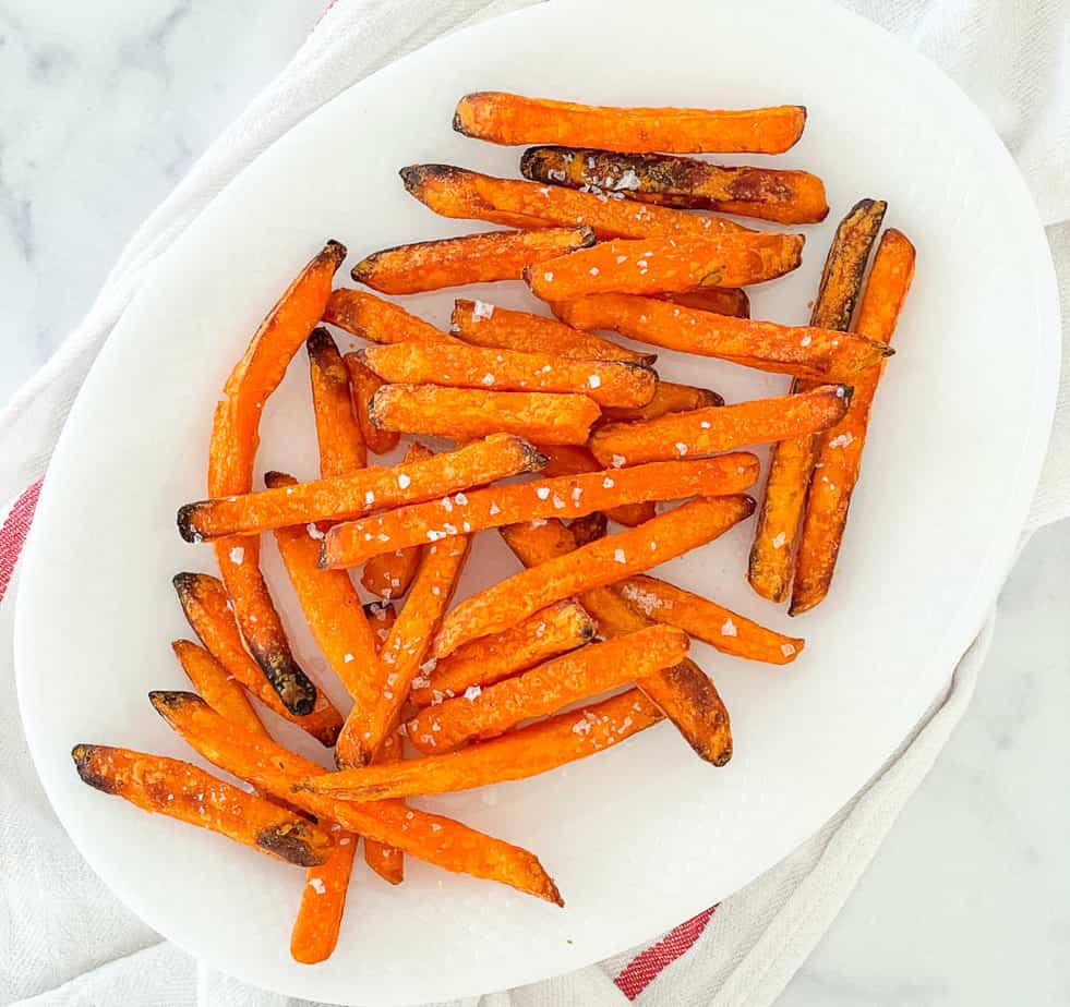 A white plate with baked sweet potato fries sprinkled with coarse salt, placed on a white surface with a red-striped towel.