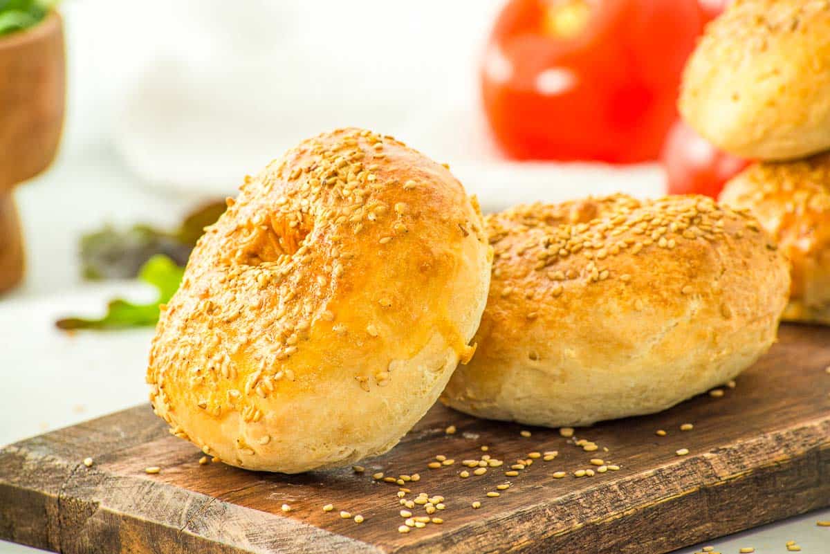 Two sesame seed bagels rest on a wooden board, with blurred tomatoes and greens in the background.